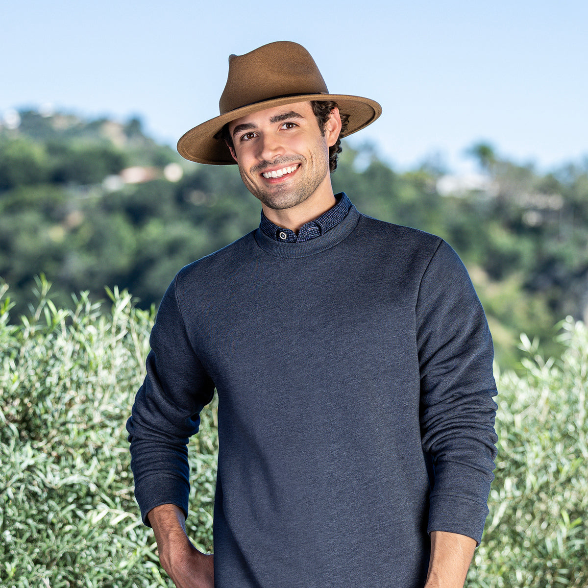 A smiling man wearing a dark heathered navy long-sleeve mock-neck shirt in the Wallaroo UPF 50+ Snowmass fedora sun hat in camel, standing outdoors in front of a blurred green landscape and blue sky.