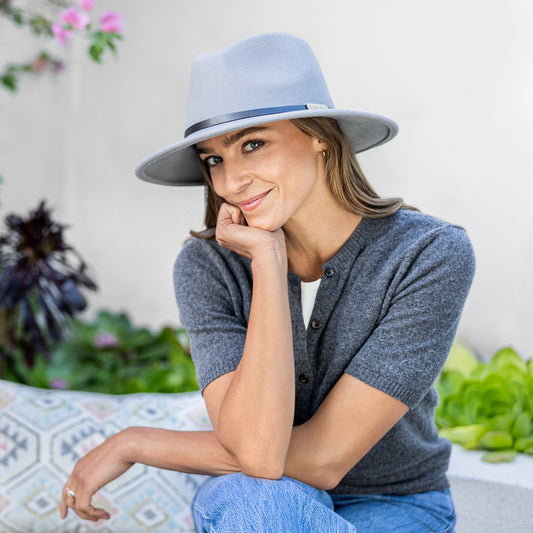 A woman wearing a gray short-sleeve button-up cardigan, blue jeans, and the gray Wallaroo UPF 50+ Snowmass fedora sun hat, sitting in a bright, modern outdoor area with plants in the background.