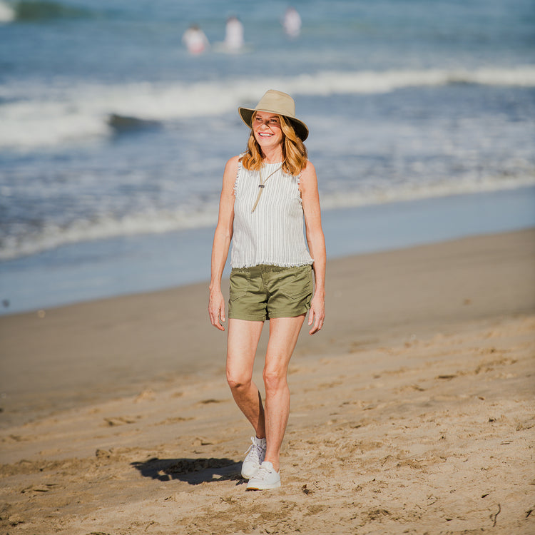 Woman enjoying walking on a beach shaded by the Wallaroo Summit Bucket Hat, designed for sunny days with wide-brim coverage and a chin strap for windy afternoons, Khaki