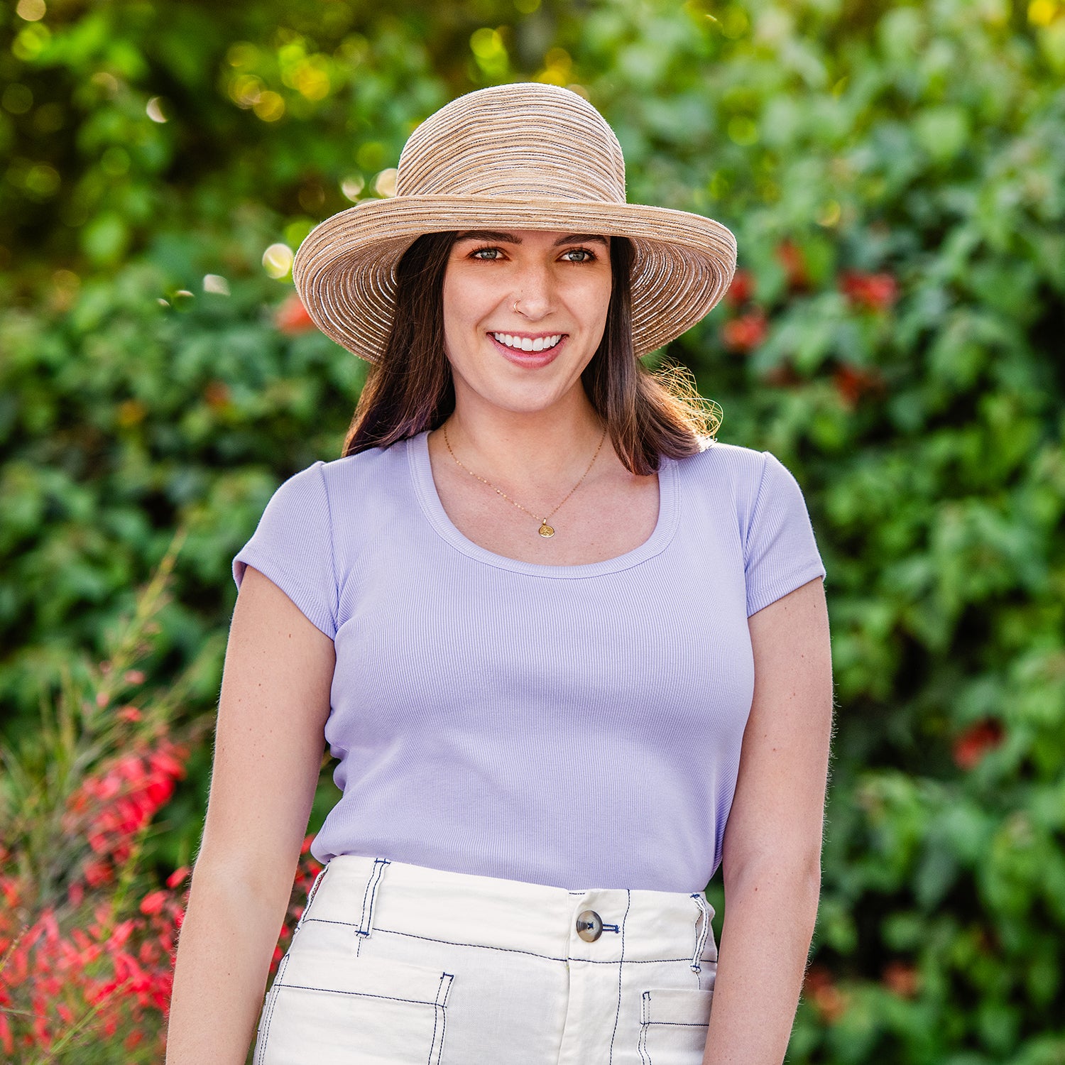 Woman wearing a Sydney hat in camel and purple shirt standing in front of green foliage