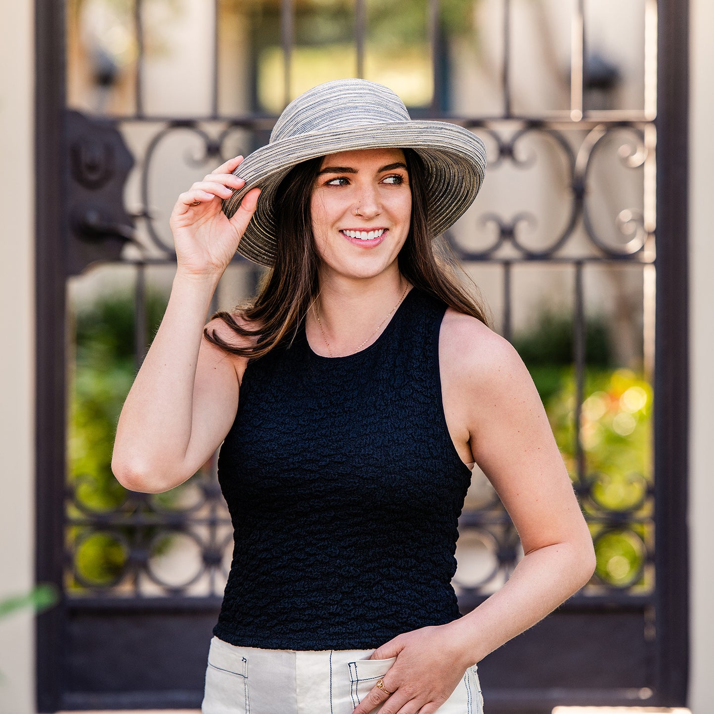 Woman wearing a Sydney wide-brimmed hat in cloud grey front of an ornate metal gate.