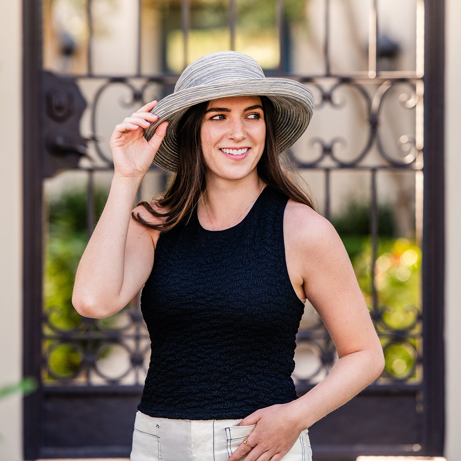 Woman wearing a Sydney wide-brimmed hat in cloud grey front of an ornate metal gate.