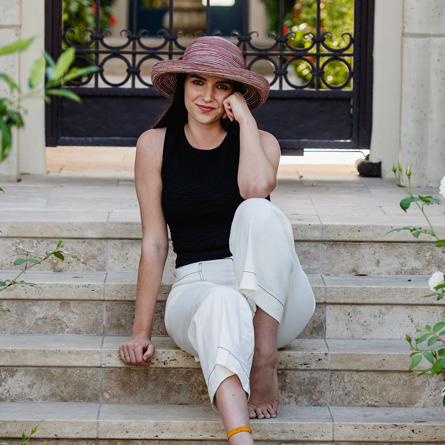Woman sitting on stone steps wearing a black tank top and white pants and Sydney sun hat, Sienna.