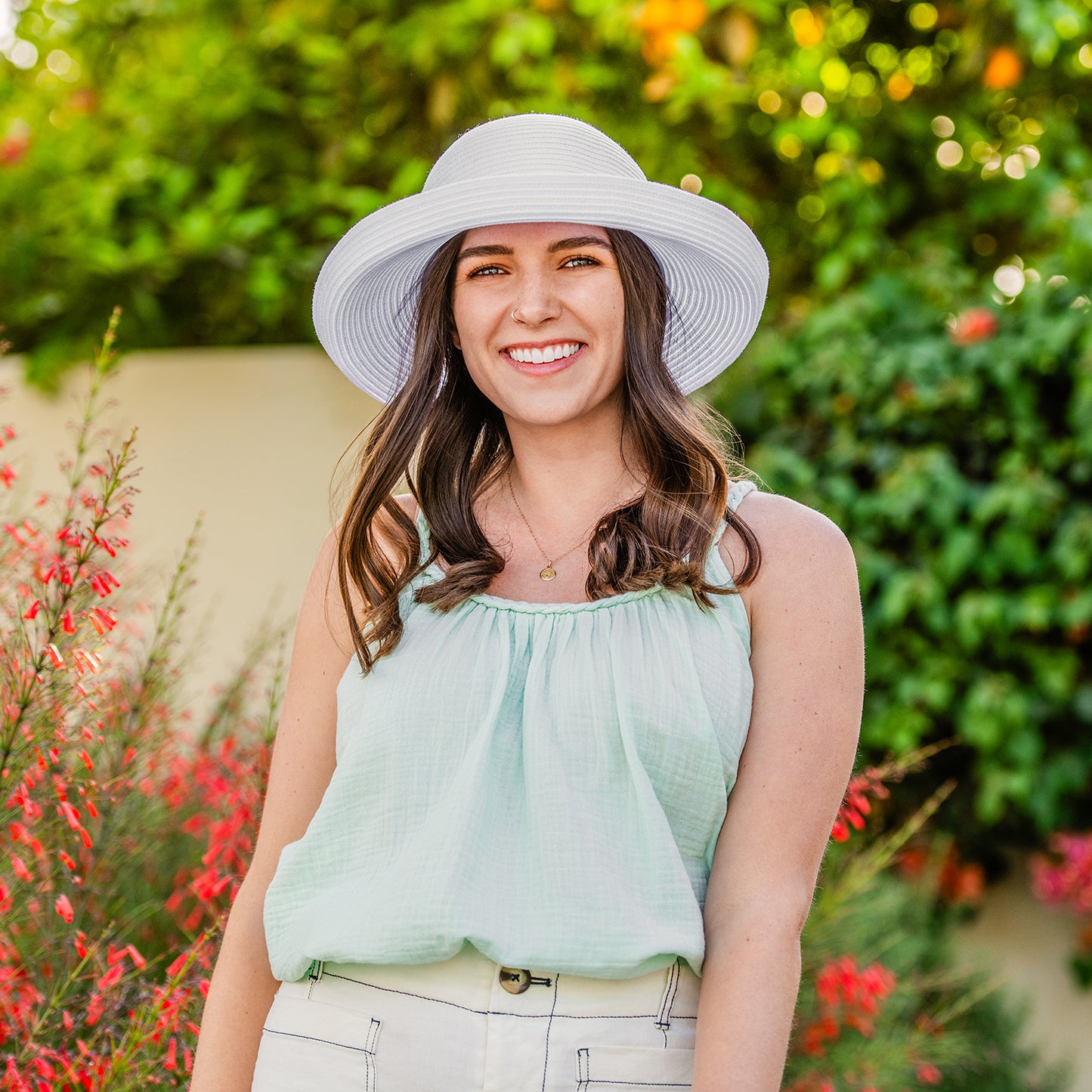 Woman wearing a light green top and white Sydney UPF 30+ sun hat in a garden setting with red flowers and green foliage.
