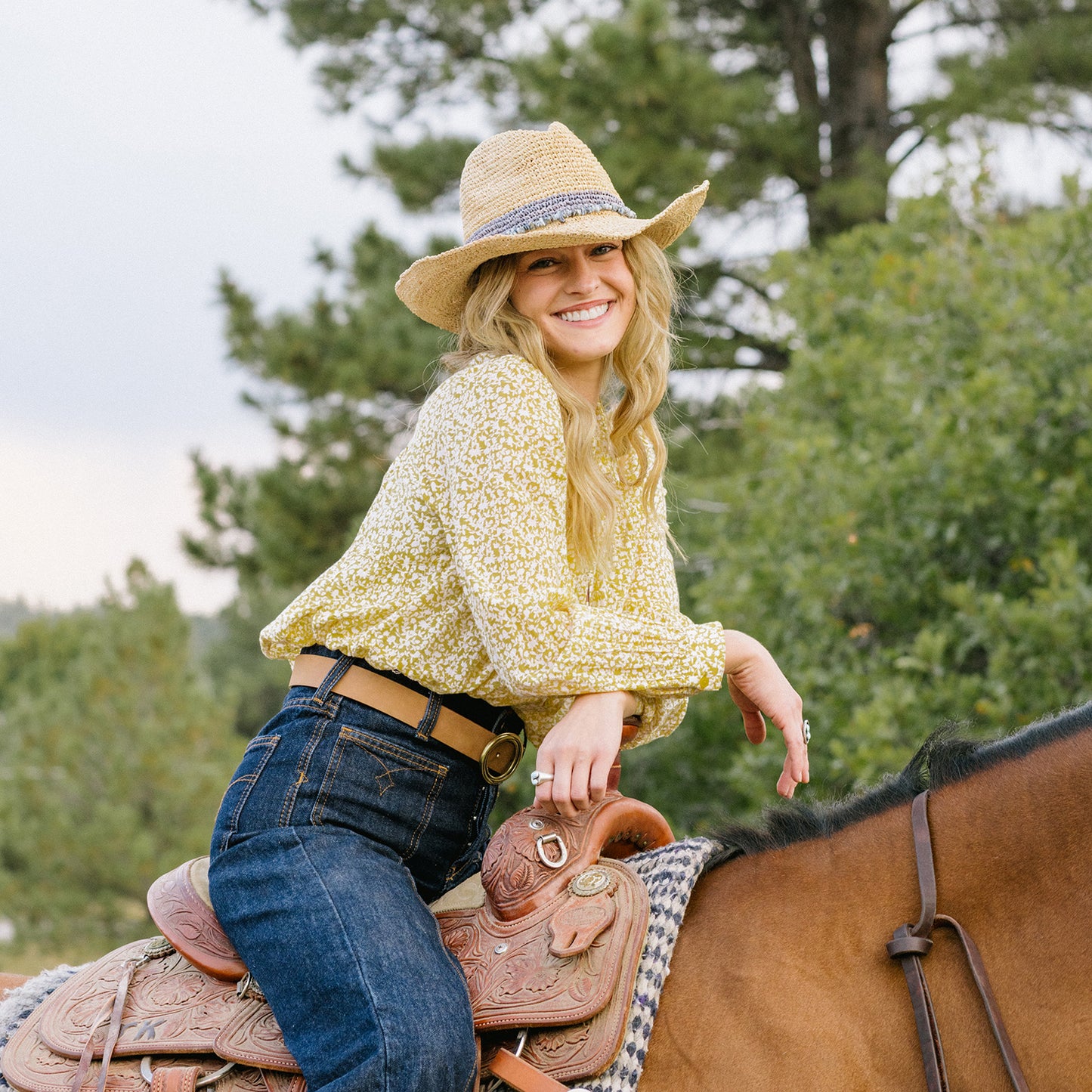 Woman wearing Tahiti Cowboy in a yellow top and blue jeans sitting on a horse with trees in the background, Dusty Blue