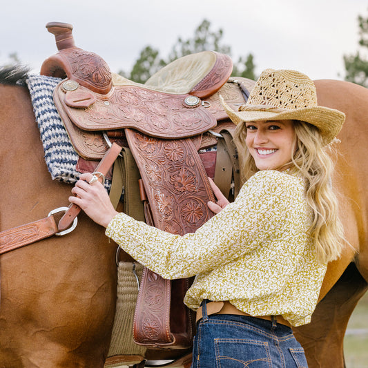 Woman in a yellow shirt and Tina raffia cowboy hat standing next to a horse with a brown leather saddle, Natural