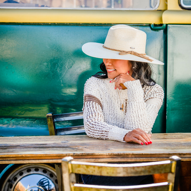 Woman lounging by a caravan at a beachside resort, staying shaded with the Wallaroo Women’s Tulum Cowgirl Hat, crafted from eco-friendly raffia straw for hot sunny days, Natural