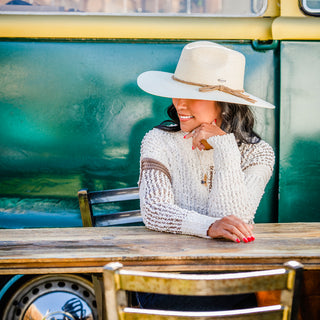 Woman lounging by a caravan at a beachside resort, staying shaded with the Wallaroo Women’s Tulum Cowgirl Hat, crafted from eco-friendly raffia straw for hot sunny days, Natural