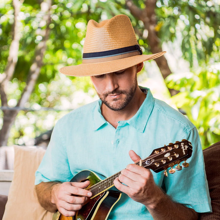 Man sitting on a resort patio with a mandolin, wearing the Wallaroo Turner Sun Hat, designed for comfort and sun protection during leisurely afternoons, Camel