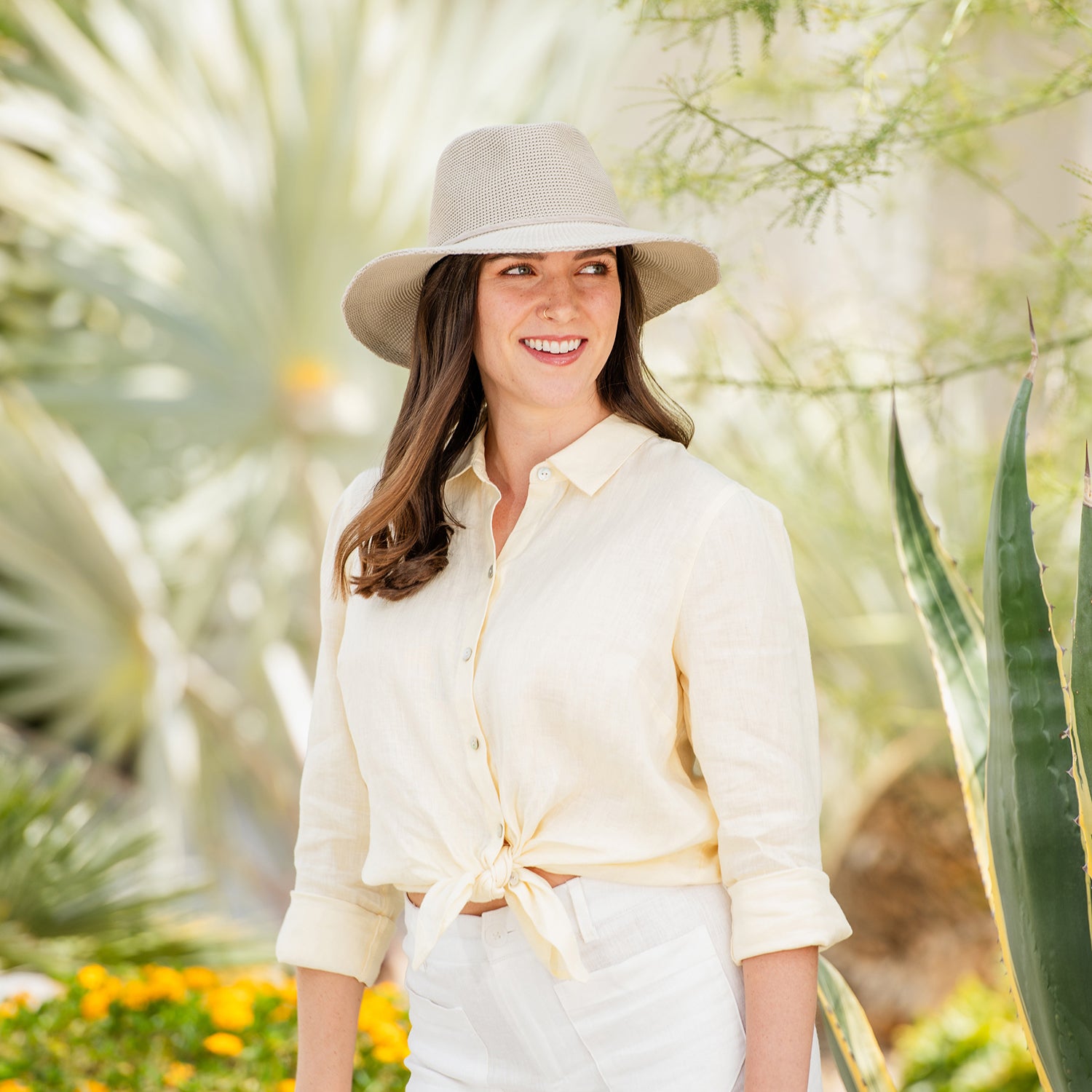 Woman wearing a light-colored shirt and Victoria Fedora sun hat standing in a garden with plants and flowers, Stone