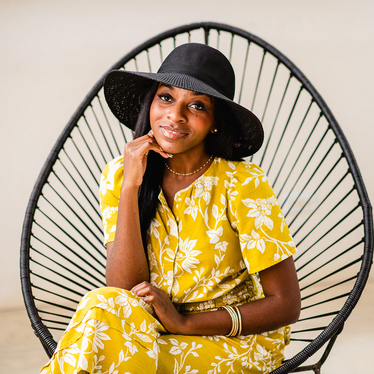 Woman sitting on a resort patio wearing the Wallaroo Victoria Diva Wide Brim Sun Hat, designed to provide sun protection with a breathable fit, Black