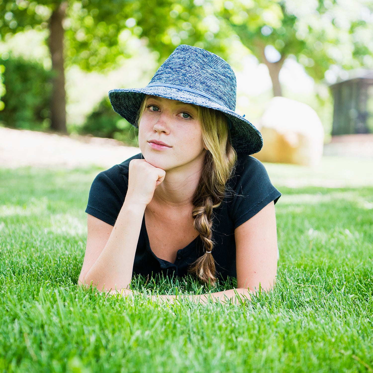STudent lying in a city park wearing the Wallaroo Women’s Victoria Fedora Sun Hat, a versatile and stylish poly-straw hat for sunny urban outings, Mixed Denim