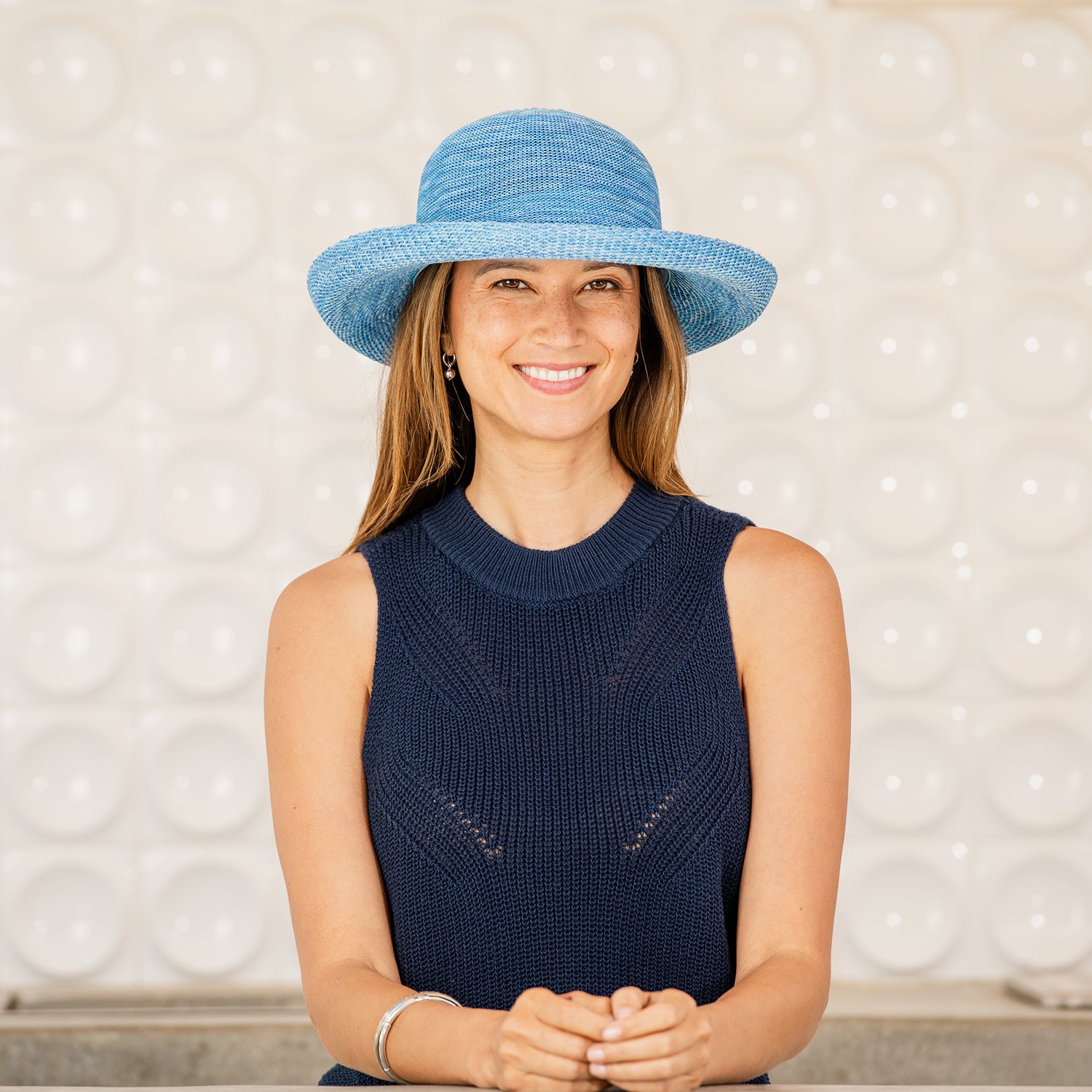 Woman wearing a Victoria sun hat and navy sleeveless top against a textured white wall, Mixed Aqua