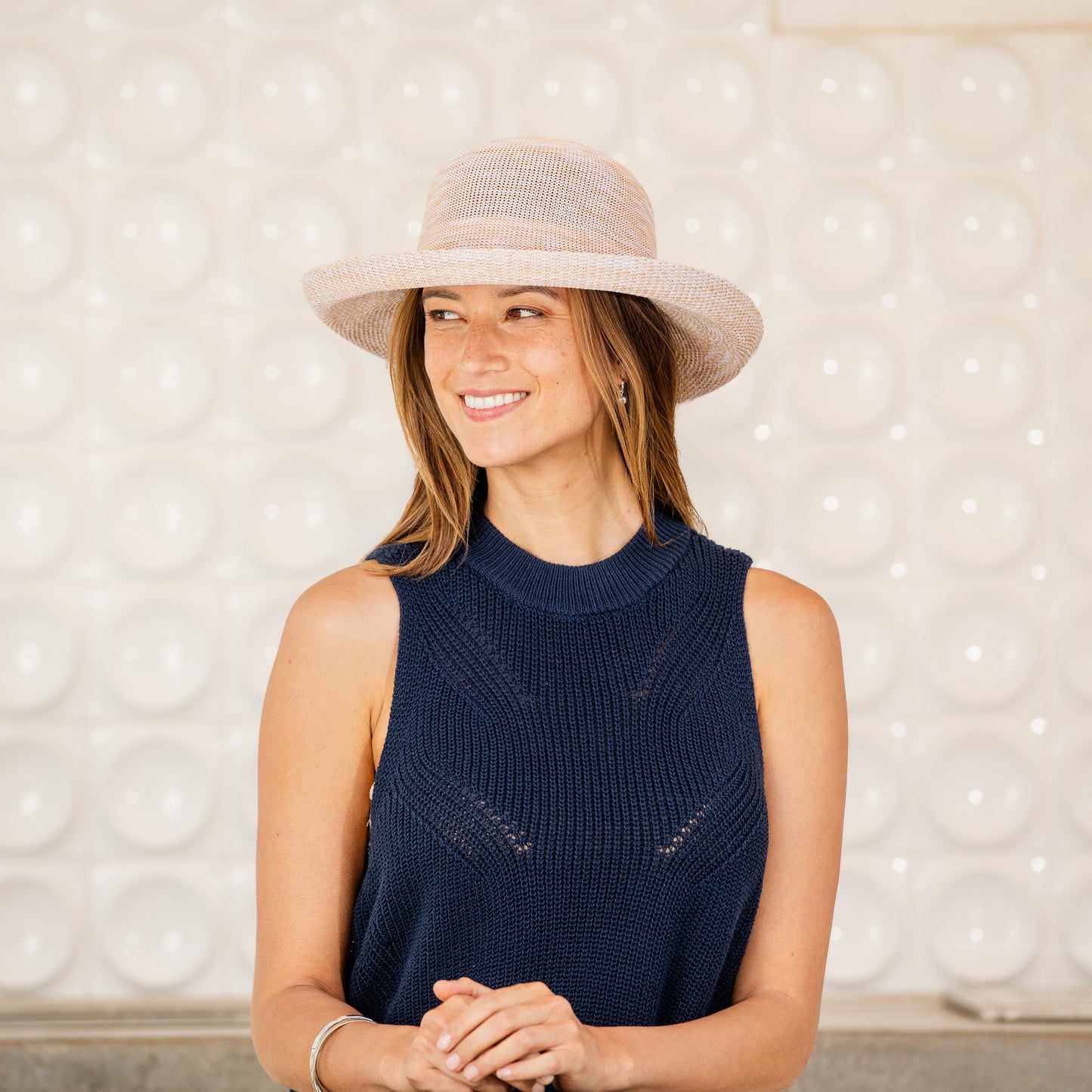 Woman wearing a navy sleeveless sweater and Victoria sun hat against a textured wall, Mixed Beige
