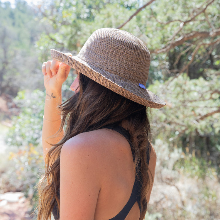 Woman walking along a scenic mountain trail, shaded by the Wallaroo Victoria Sun Hat, offering breathable sun protection for outdoor adventures, 'Mixed Camel'