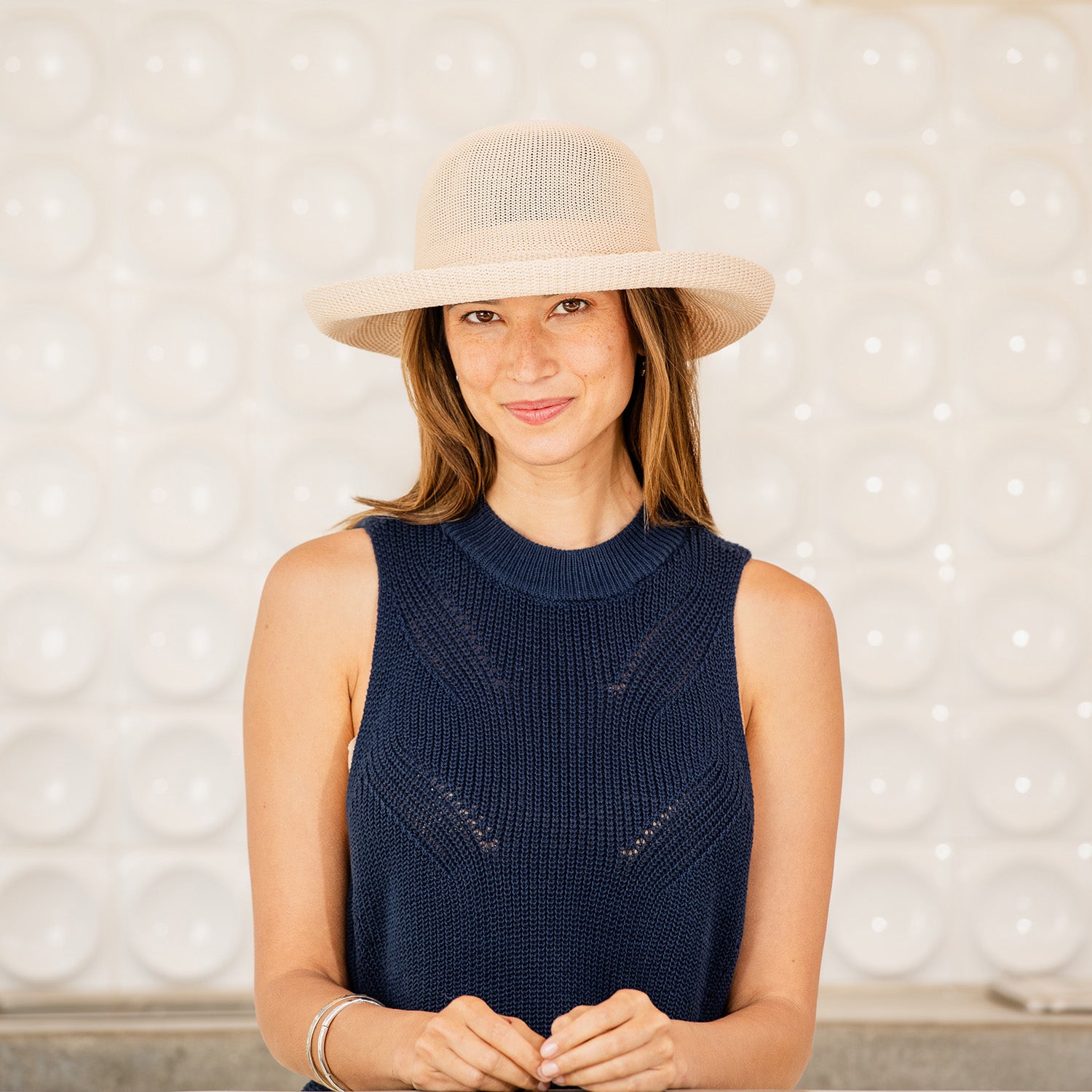 Woman wearing a navy sleeveless top and Victoria sun hat against a textured wall, Tan