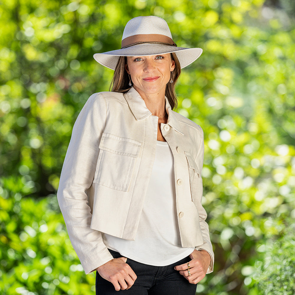 A woman wearing the Cypress UPF 50+ packable wide-brimmed light mixed camel hat, with a light beige jacket over a white top, standing outdoors against a bright, blurred green foliage background.