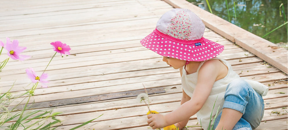 Toddler wearing a pink polka dot Platypus sun hat on a wooden dock with flowers.